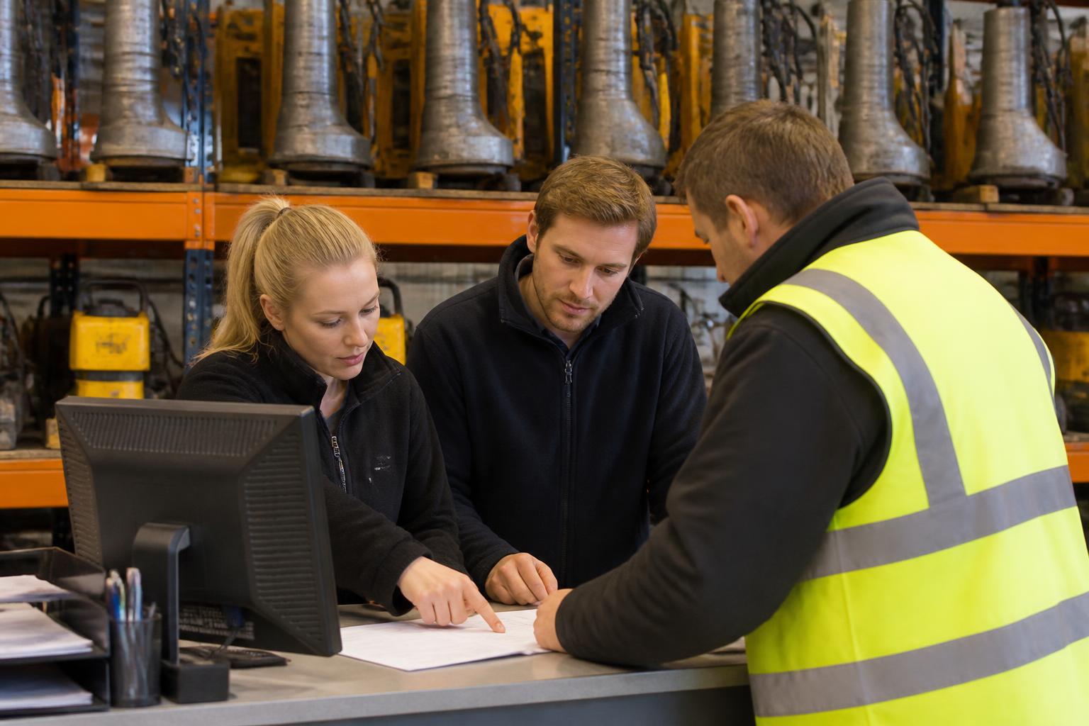 Plant Hire Insurance 2 Hire desk staff going through paperwork with a site manager at a busy UK plant and tool hire counter, with breakers and compactors racked behind them
