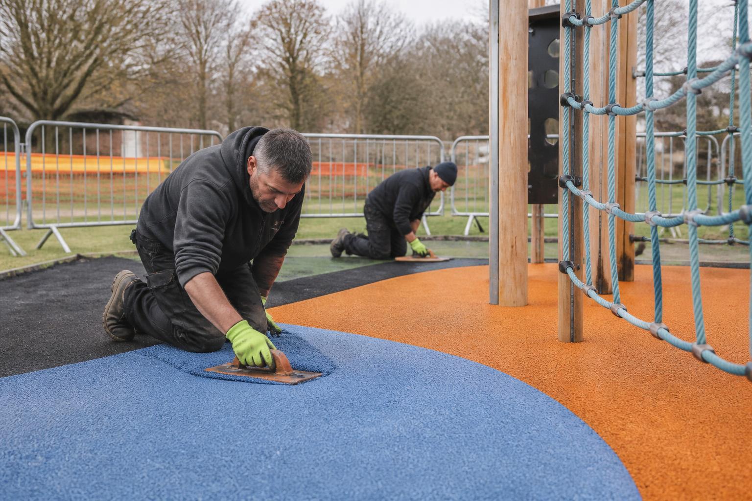 Sports Surfacing Insurance 2 Two installers laying colourful wet pour safety surfacing around new climbing equipment in a UK park playground, temporary barriers set around the site